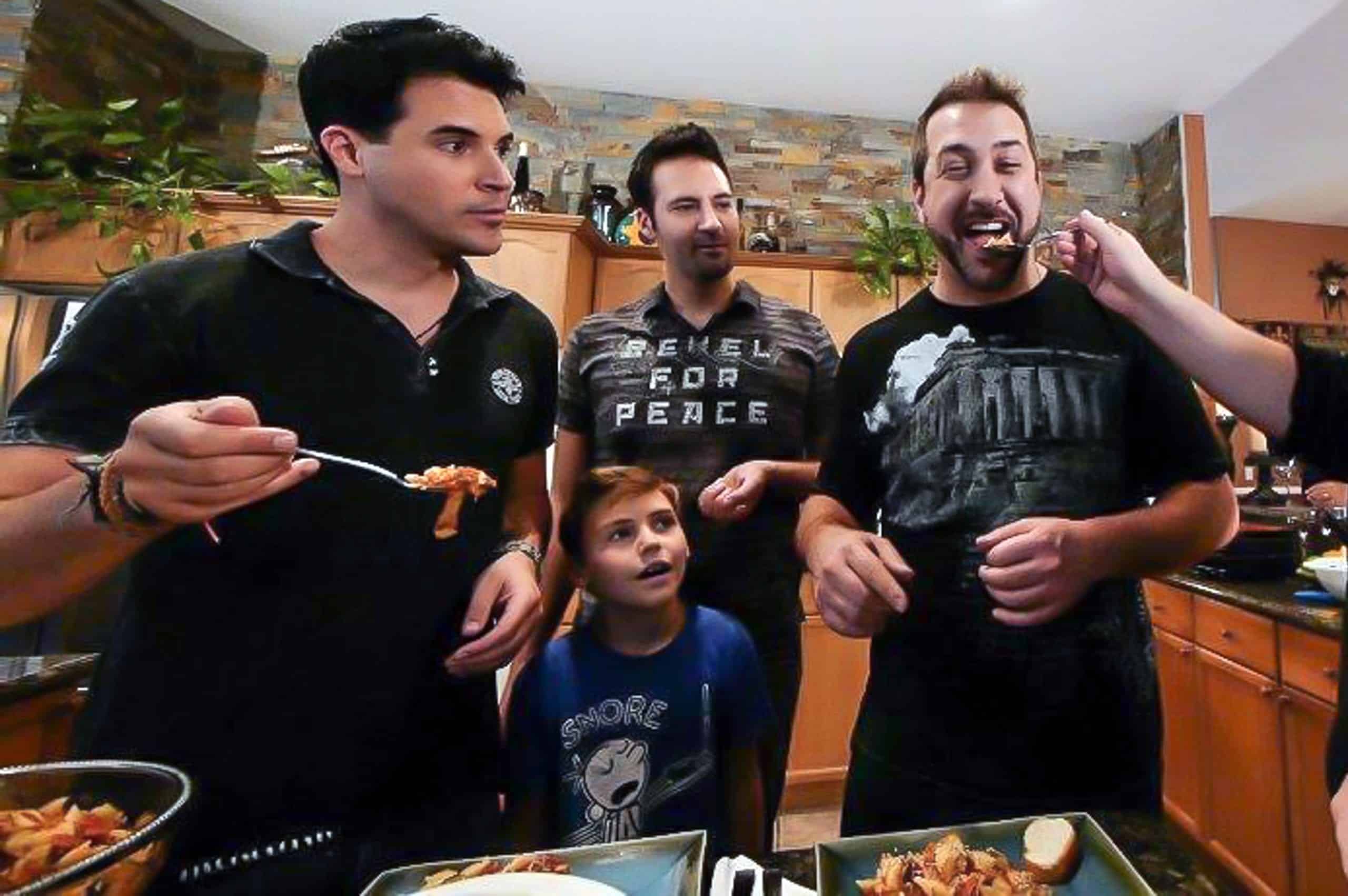 Three men and a boy in a kitchen, with one man holding a fork of food and another being fed.