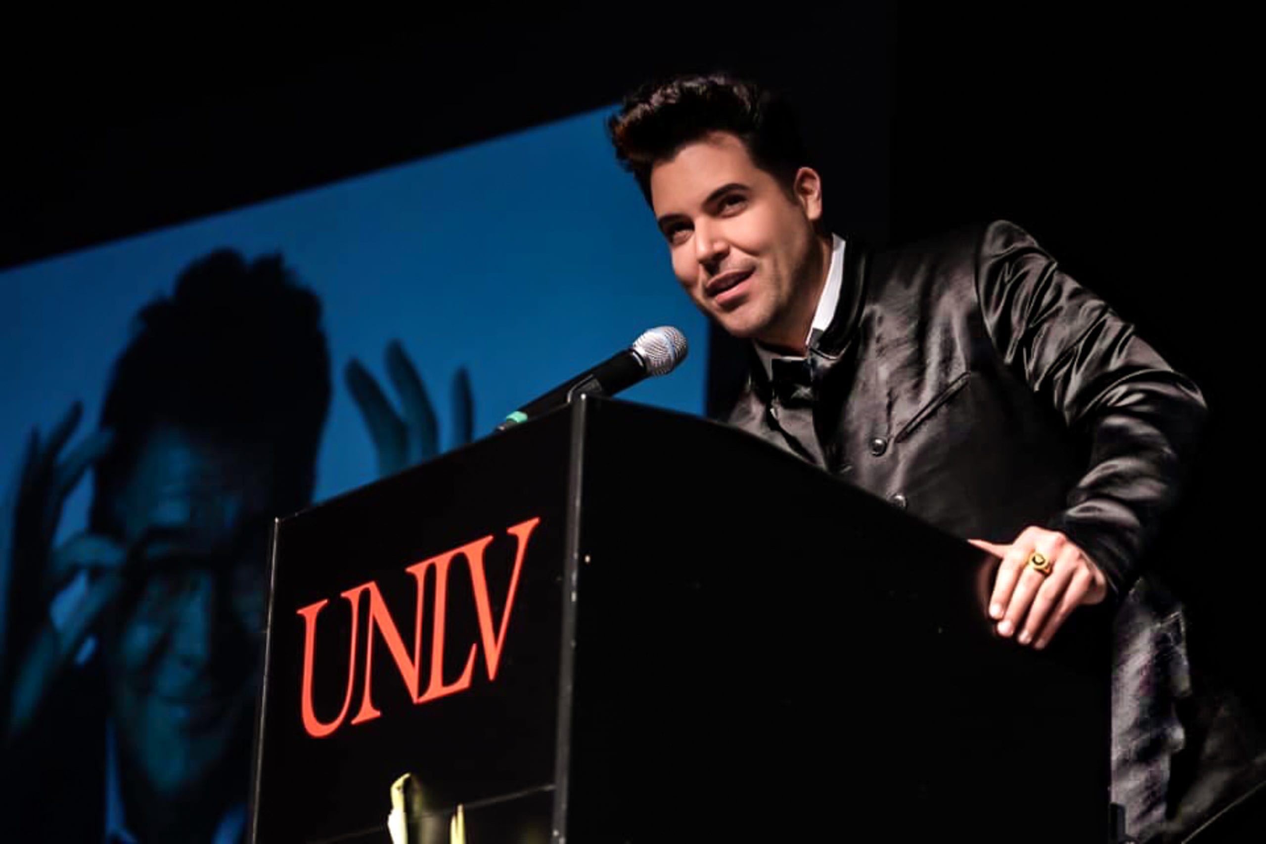 A man in a black satin jacket speaks at a podium with "UNLV" in red text, a screen behind him.