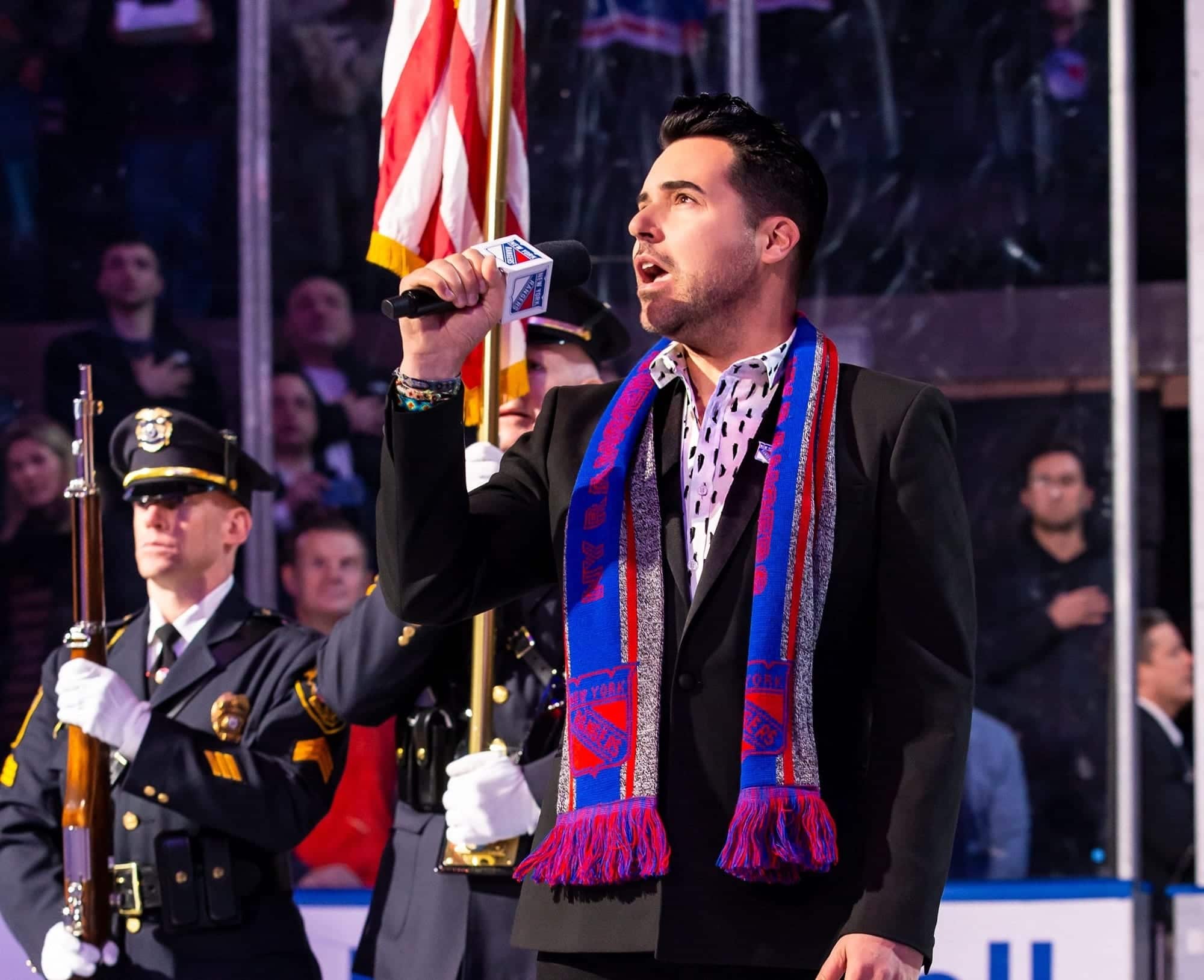 A man with a beard and a blue and red scarf sings into a microphone, with a police officer holding a rifle and an American flag behind him.