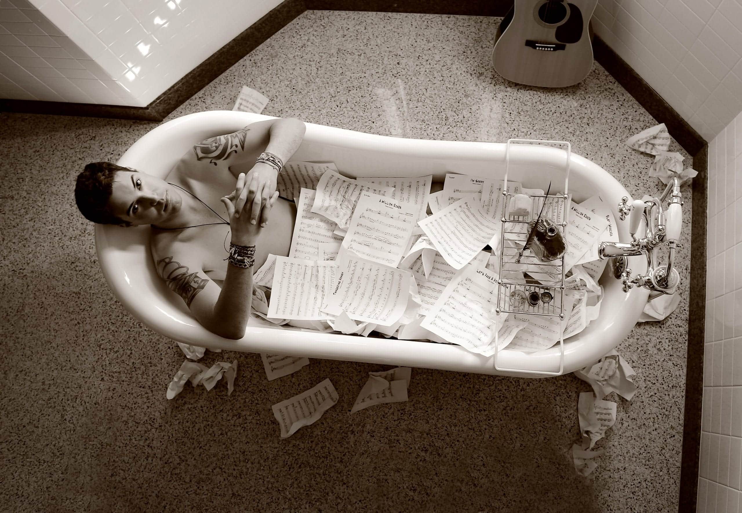 A person with tattoos and bracelets lies in a bathtub filled with sheet music, with a guitar nearby.