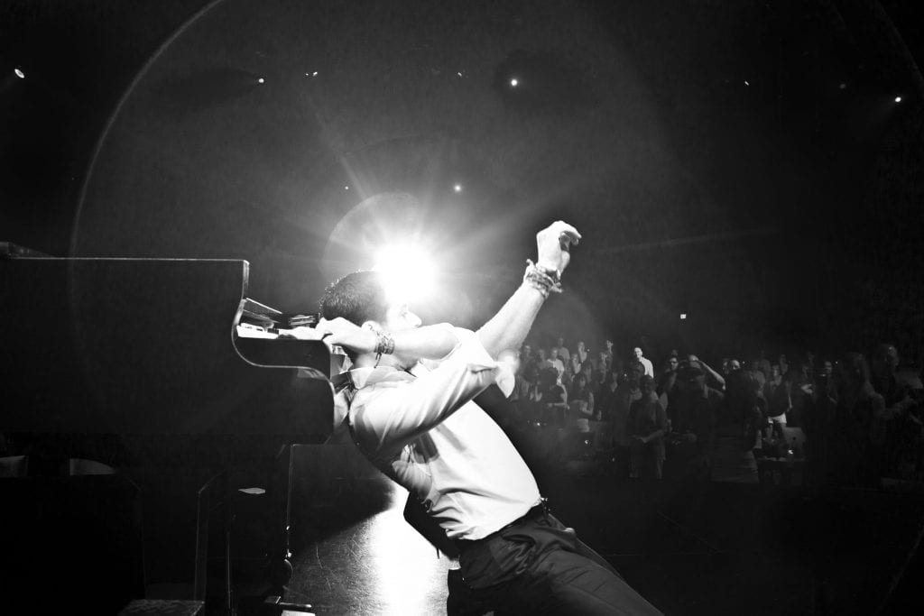 A man in a white shirt and dark pants leans back from a piano, arm raised, bathed in stage light.