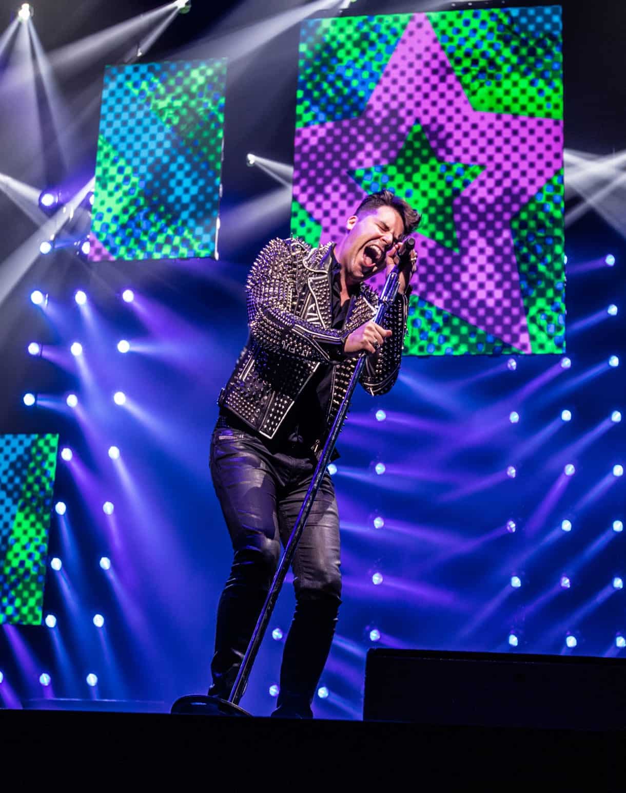 A male singer in a studded jacket screams into a microphone on stage with colorful star screens.