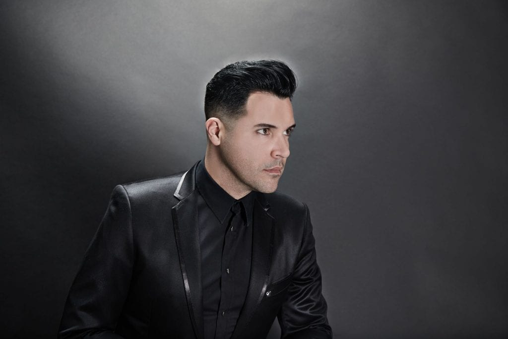 A man with dark hair in a pompadour, wearing a black suit and shirt, looks right against a gray background.