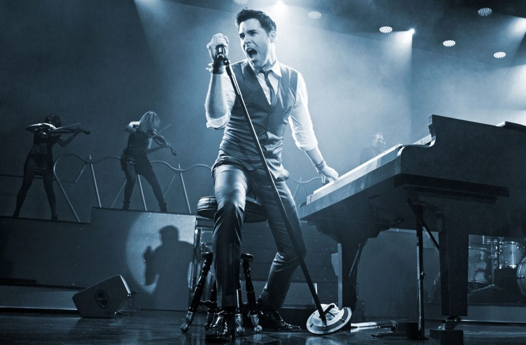 A male singer in a vest and tie sings into a microphone on stage next to a piano, with violinists in the background.