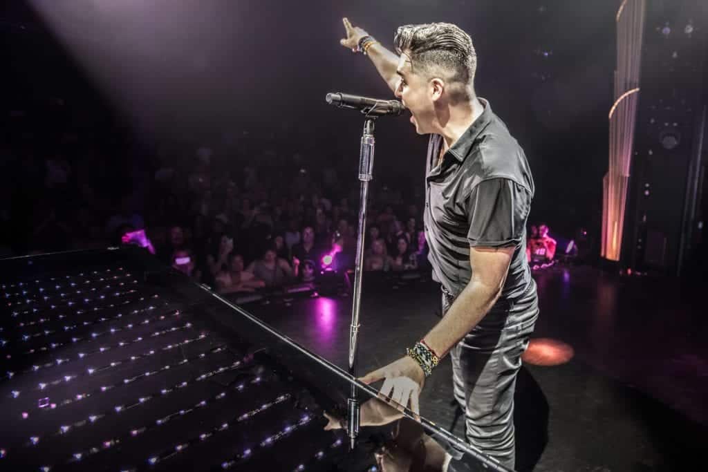 A male singer in a black shirt points upwards while singing into a microphone on a stage with a crowd.