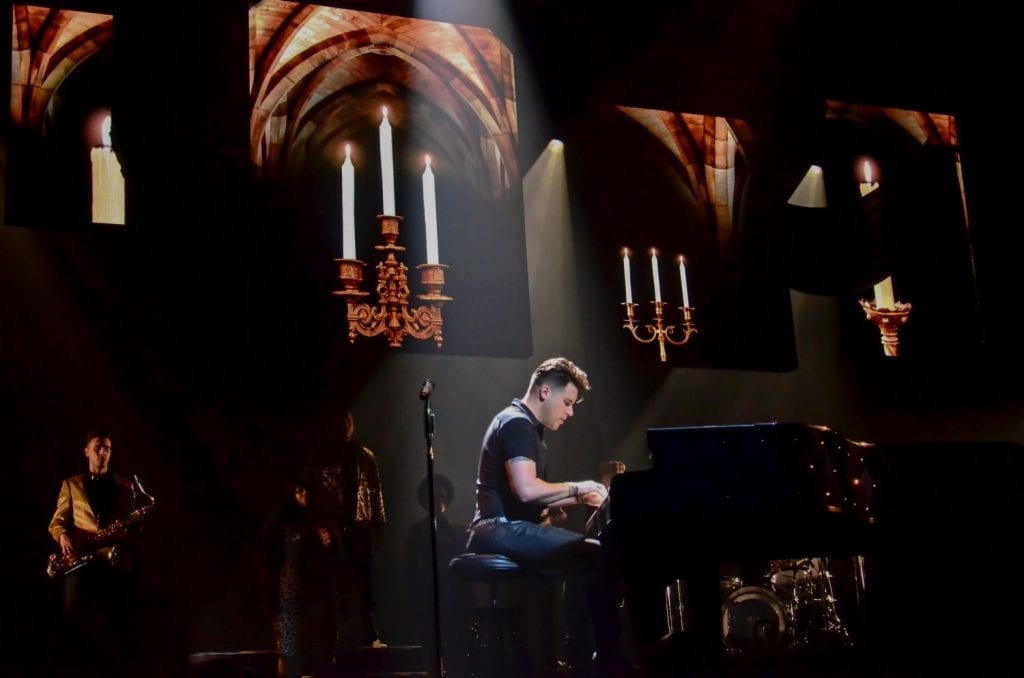 A man in a black shirt plays a grand piano on a dark stage with projected images of candles and arches.