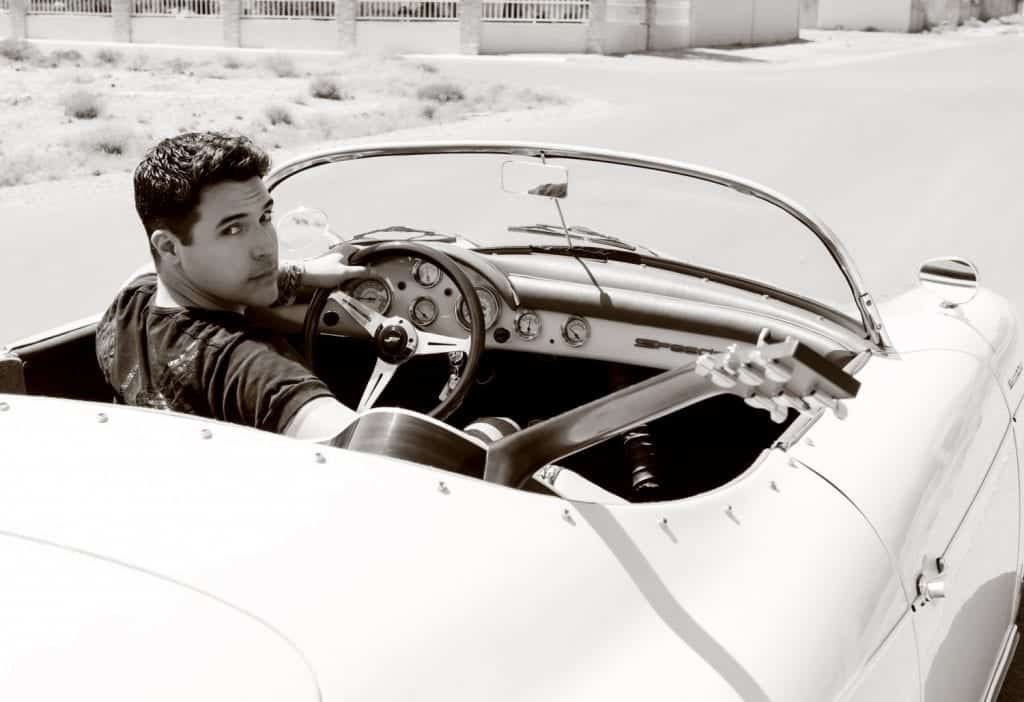 A young man in a black shirt sits in a vintage convertible with a guitar, looking over his shoulder.