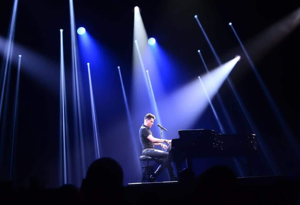 A man in a black shirt plays a grand piano on a dark stage illuminated by blue and white spotlights.