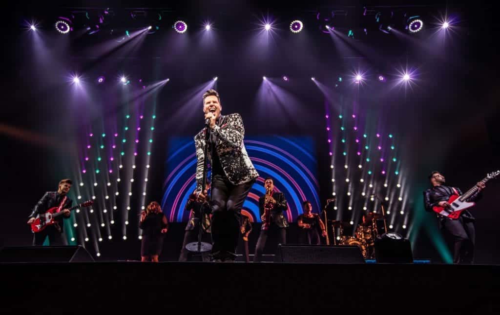A male singer in a patterned jacket performs on stage with a band under purple and teal lights.
