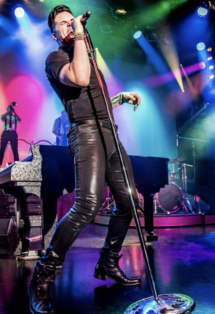 A male singer in a black shirt and shiny black pants sings into a microphone on a colorful stage.