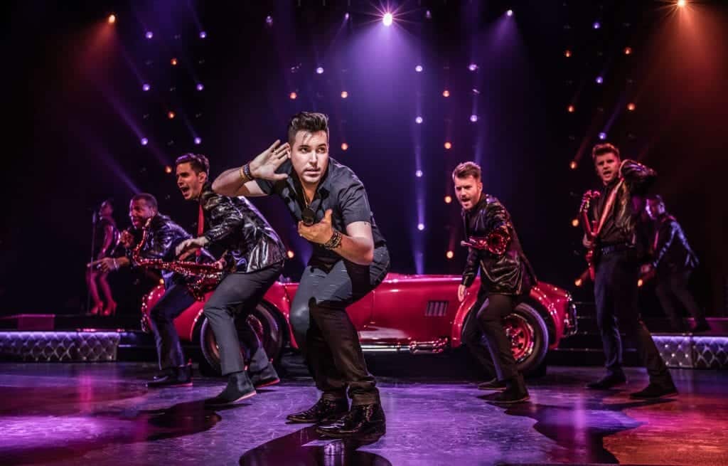 A male singer in a black shirt holds a mic, gesturing to his ear, with musicians and a red car behind him.