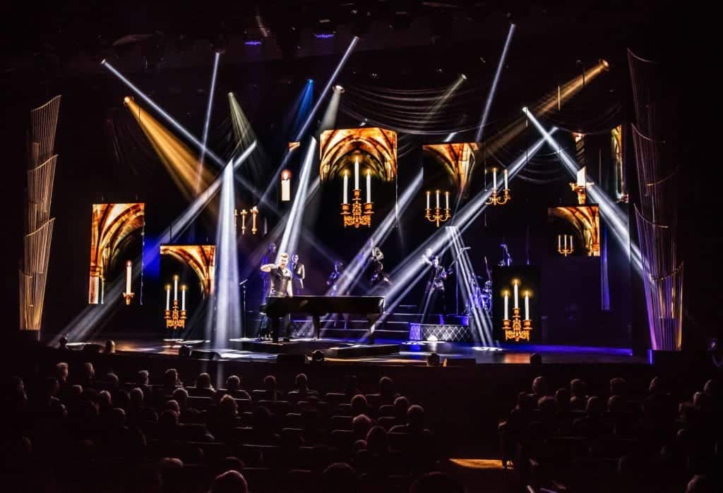 A performer stands by a grand piano on a stage with bright spotlights and screens displaying chandeliers.