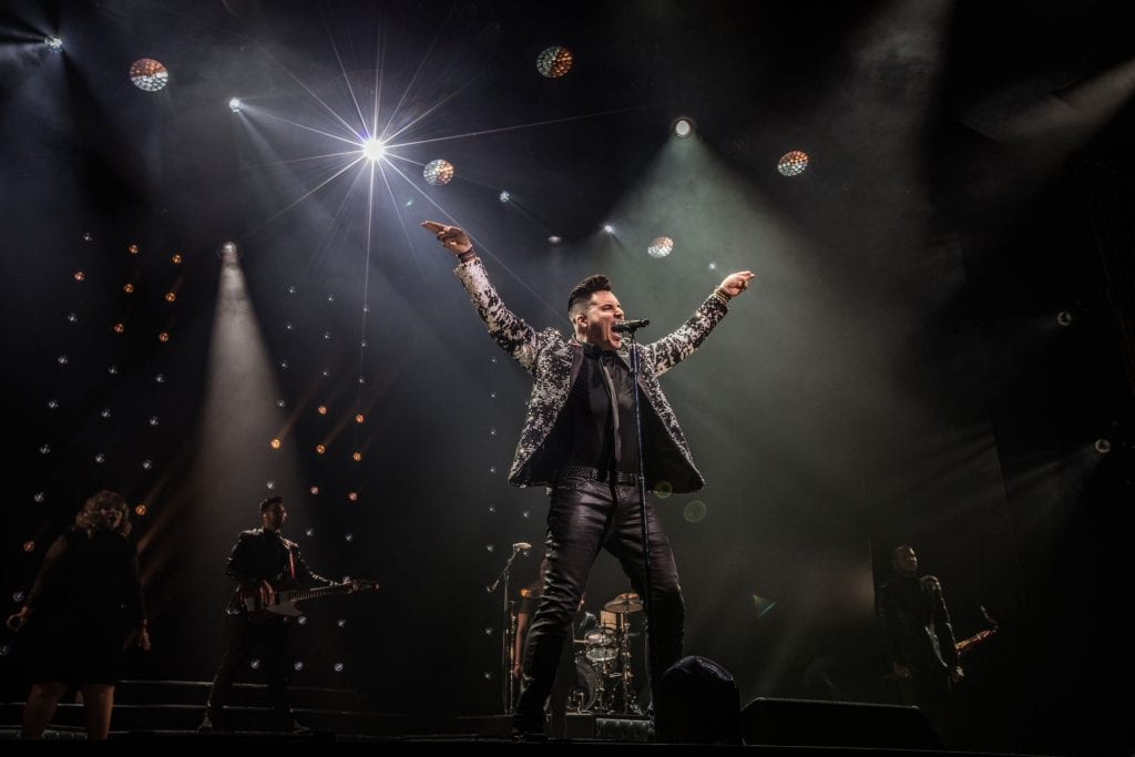 A male singer in a patterned jacket with arms raised, performing on a dark stage with bright spotlights.