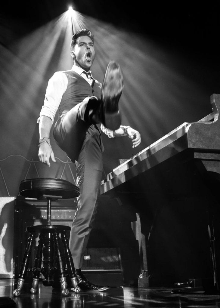 A man in a vest and dress shirt, mouth open, kicks his leg up next to a piano.