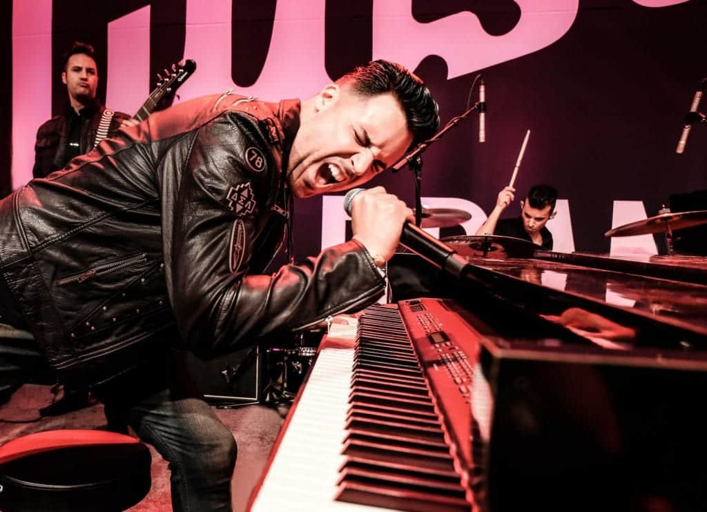 A man in a black leather jacket sings into a microphone while playing a keyboard on a stage.
