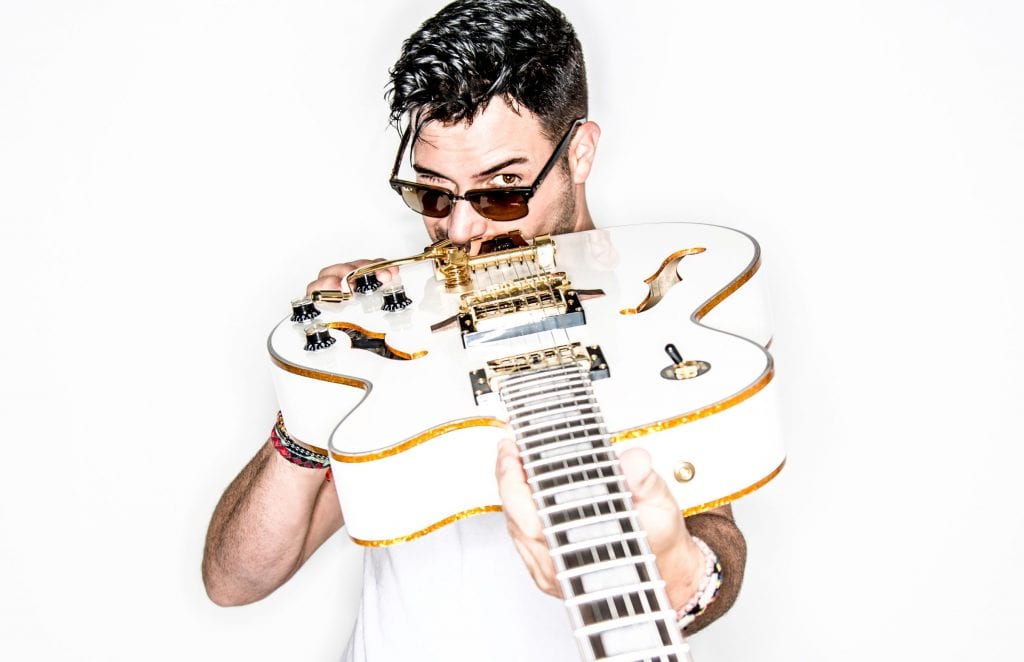 A man with dark hair and sunglasses holds a white electric guitar with gold hardware, looking over the top.
