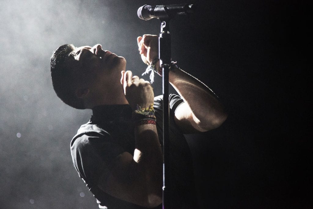 A man with dark hair and a black shirt looks up, bathed in bright stage light, with a microphone stand in front of him.