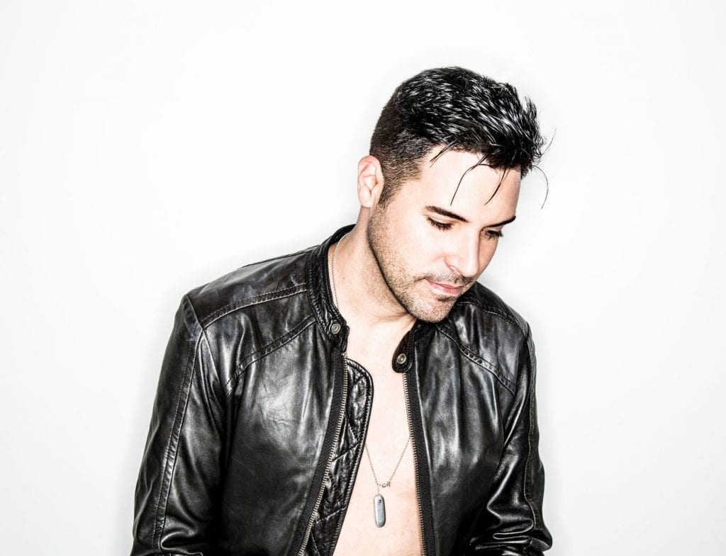 A man with dark hair and stubble wears an open black leather jacket and a pendant necklace.
