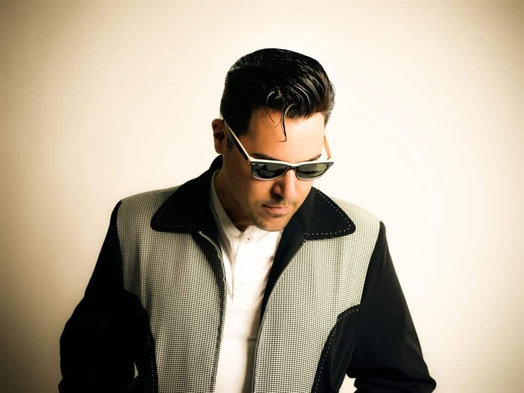 A man with slicked-back dark hair and white-framed sunglasses looks down, wearing a black and white jacket.