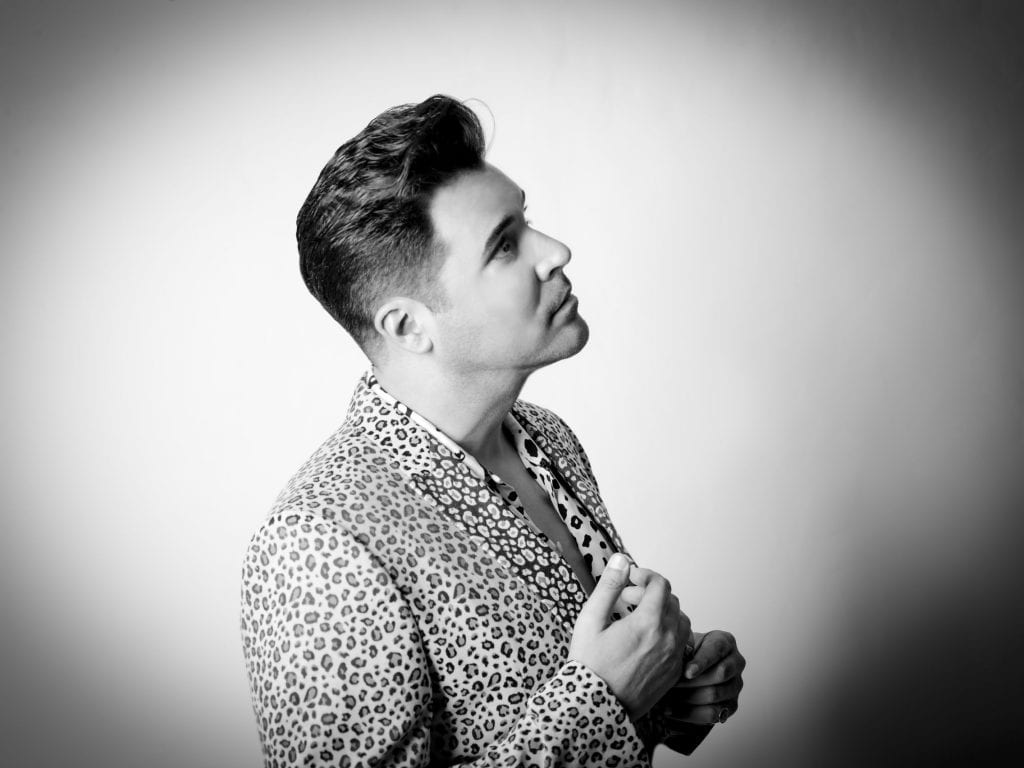A man with a pompadour hairstyle and a leopard print jacket looks up and to the right in black and white.