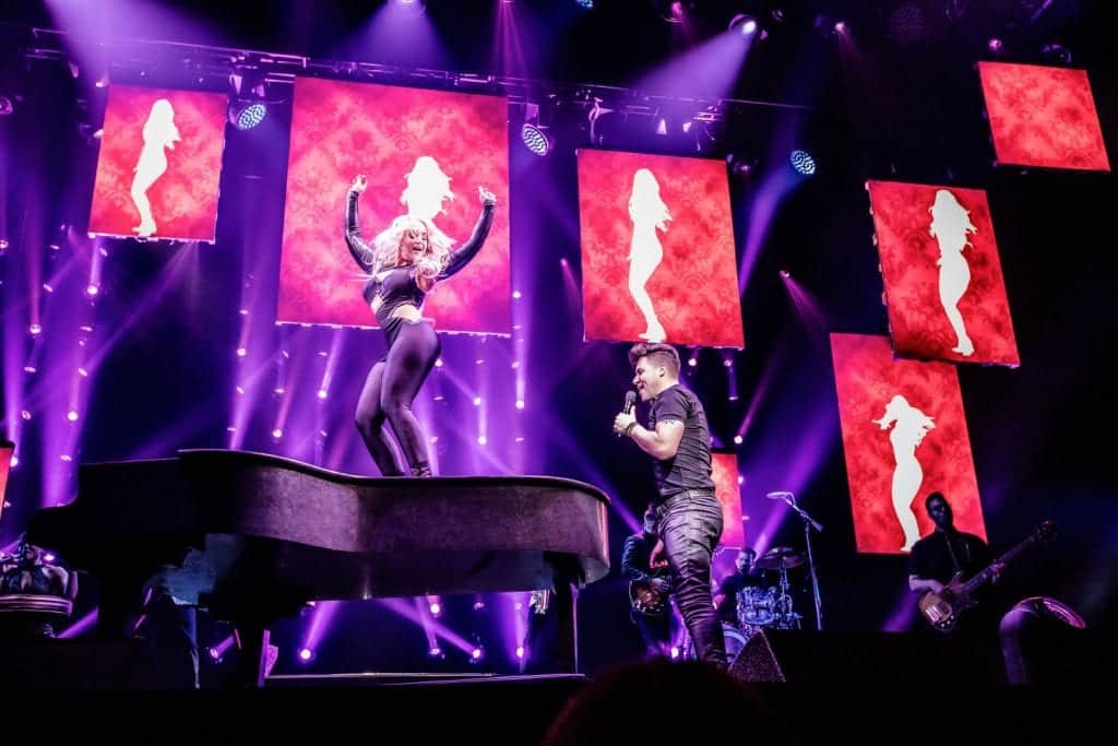 A female singer in black performs on a grand piano, while a male singer with a microphone stands beside it.