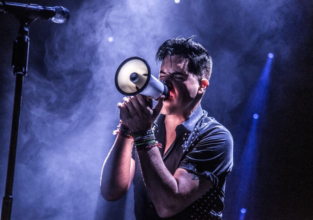 A man with dark hair and a black shirt sings into a white megaphone on a smoky stage.