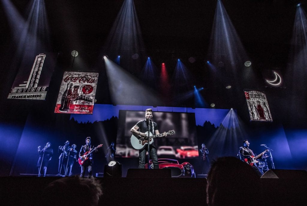 A male singer in black performs on stage with a band, surrounded by spotlights and vintage posters.