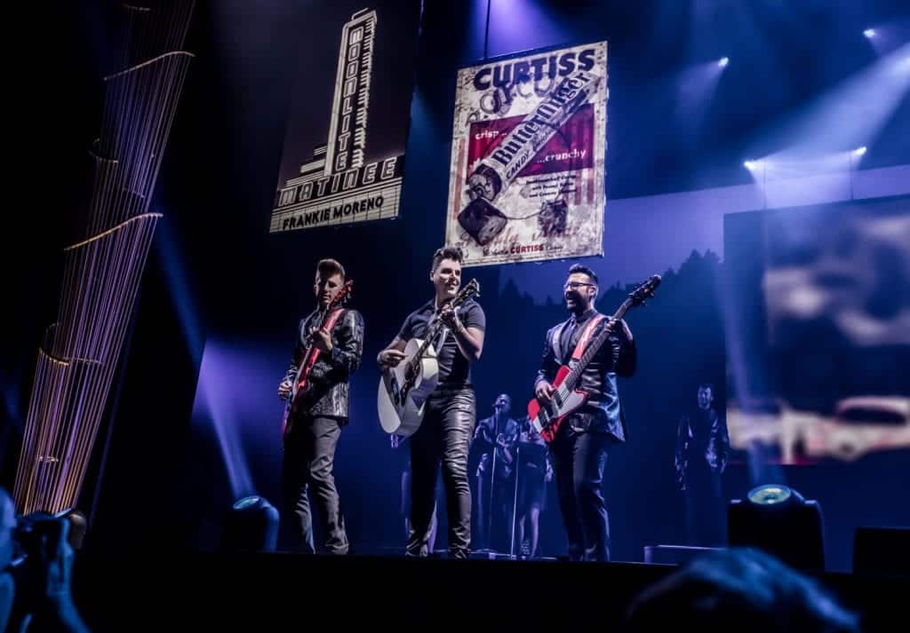 Three musicians play guitars on a dark stage under spotlights and two large banners, one reading "CURTISS Butterfinger."