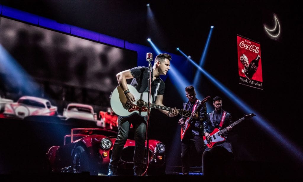 A band plays guitars on a dark stage with a red "Coca-Cola" sign and classic cars in the background.