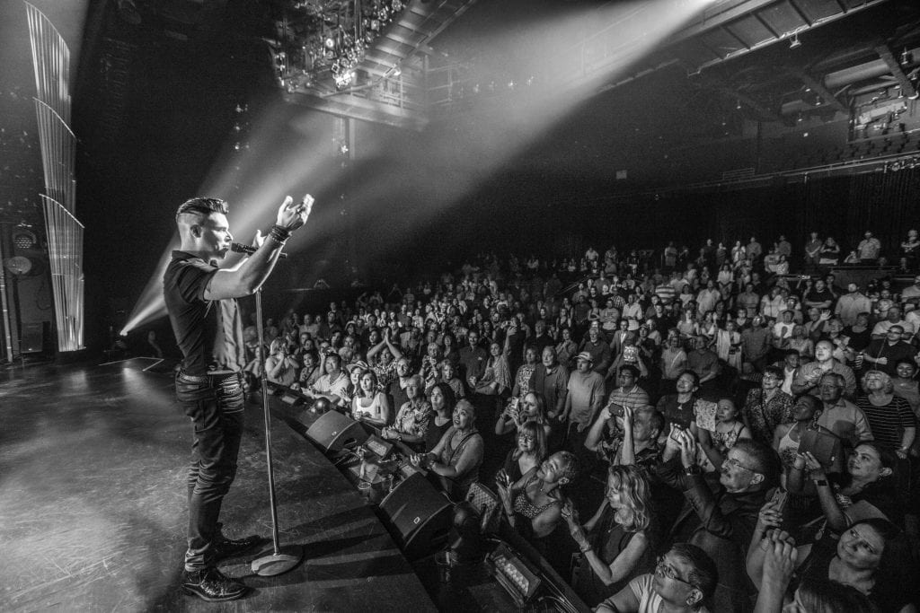 A male performer with a microphone stands on stage, facing a large, excited audience in a dark venue.