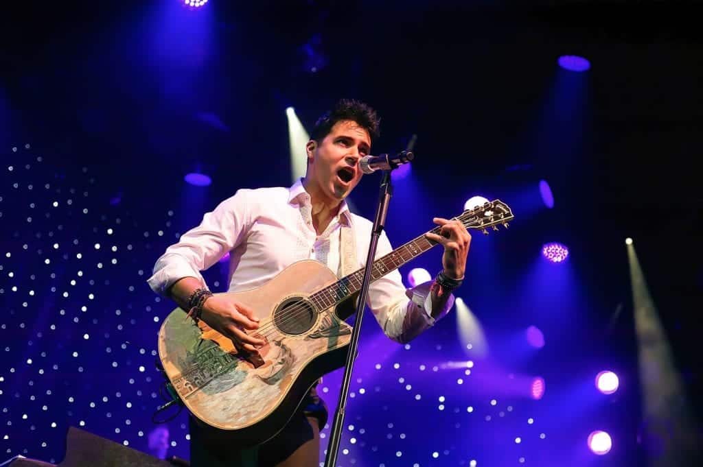 A male singer in a white shirt plays an acoustic guitar with a painted scene on stage under blue lights.