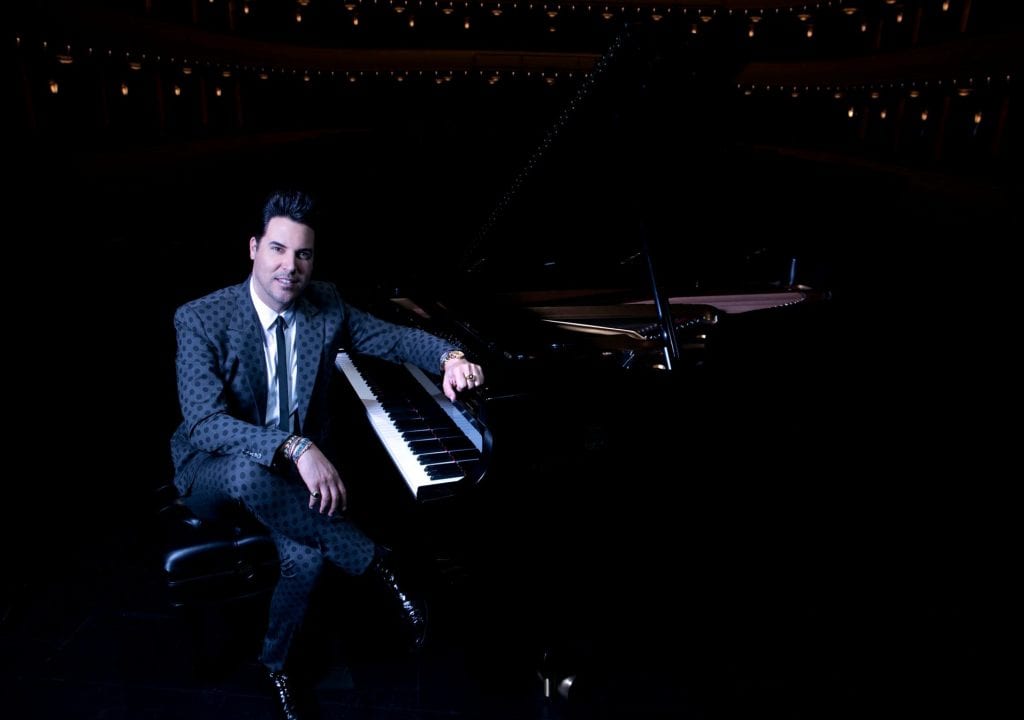 A smiling man in a black polka-dotted suit sits on a piano bench, leaning on a grand piano.