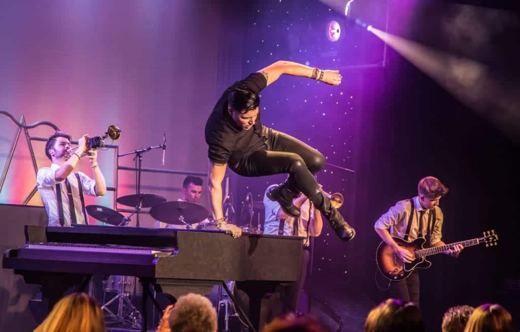A male singer in black leather jumps mid-air over a piano, surrounded by musicians on a purple-lit stage.