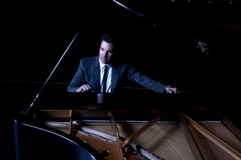 A man in a dark patterned suit and tie sits at a grand piano, looking to the left.