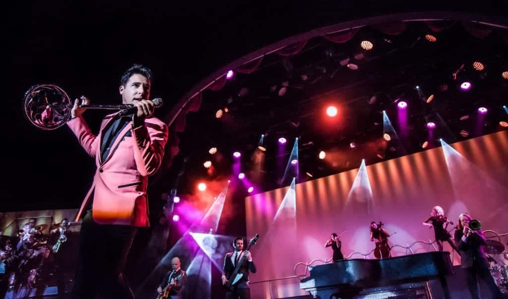 A man in a pink suit sings into a microphone while holding a banjo, with a band on stage below.
