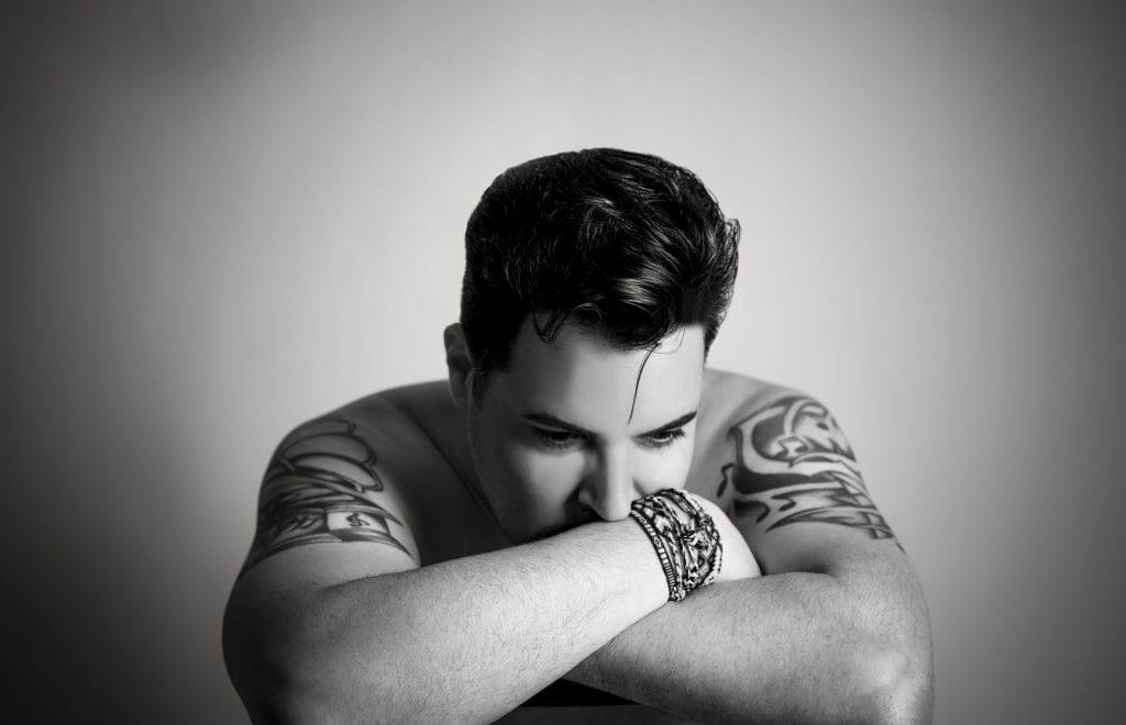 A shirtless man with dark hair and tattoos on both arms rests his chin on his forearm, looking down.