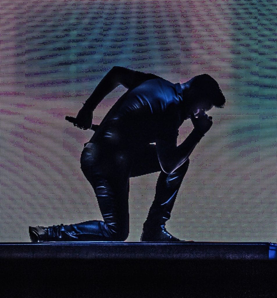 A dark silhouette of a person in a shiny outfit kneels on a stage, holding a microphone.