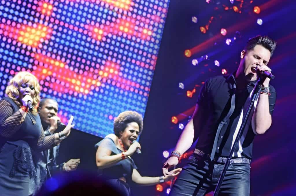A male singer in a black shirt and leather pants sings into a microphone with backup singers on a stage.