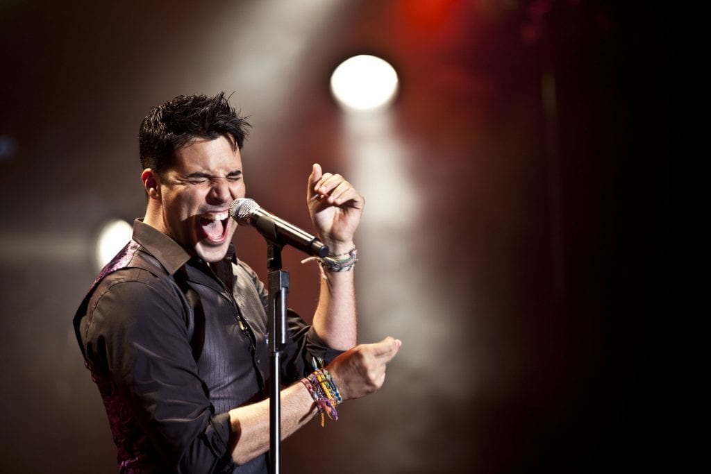 A male singer with spiked hair and a black shirt passionately sings into a microphone on a dark stage.
