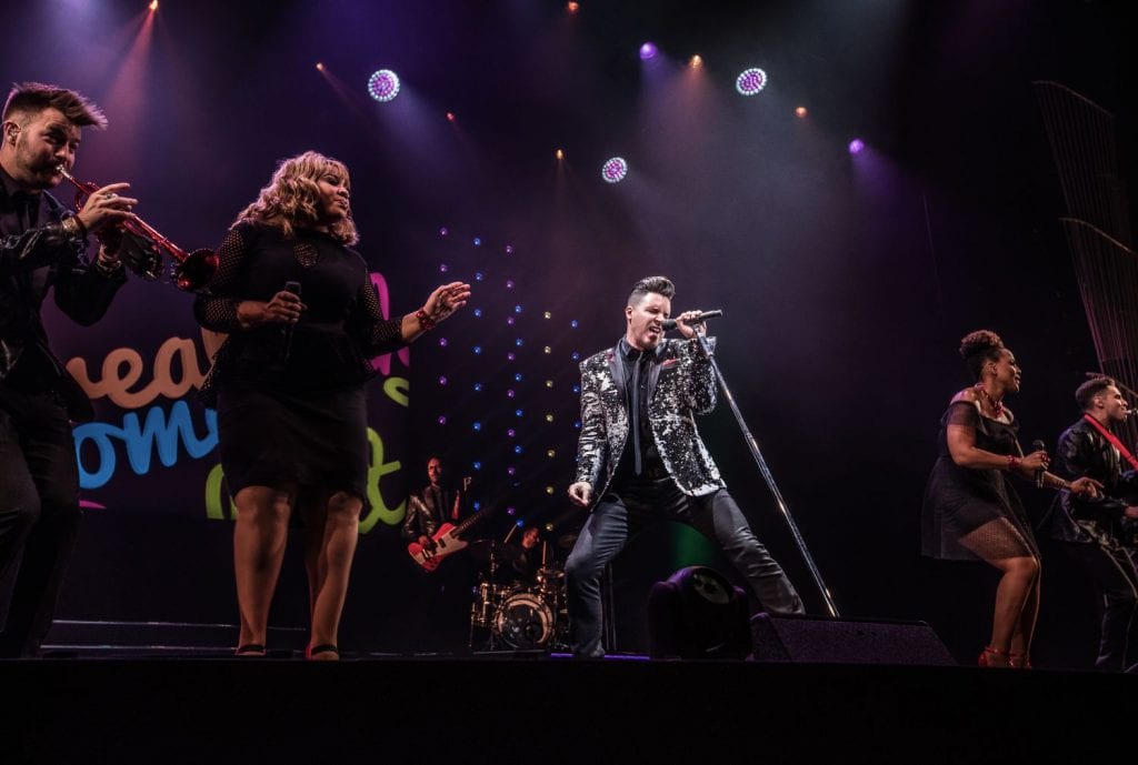 A band performs on stage with a male singer in a sequined jacket and two female backup singers.