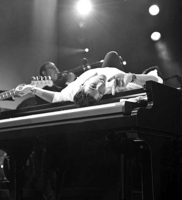 A man in a white shirt lies on his back across a piano, looking up at the camera.