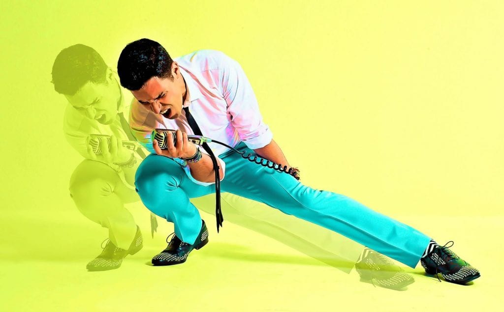 A man in a white shirt and turquoise pants sings into a microphone, lunging forward with an open mouth.