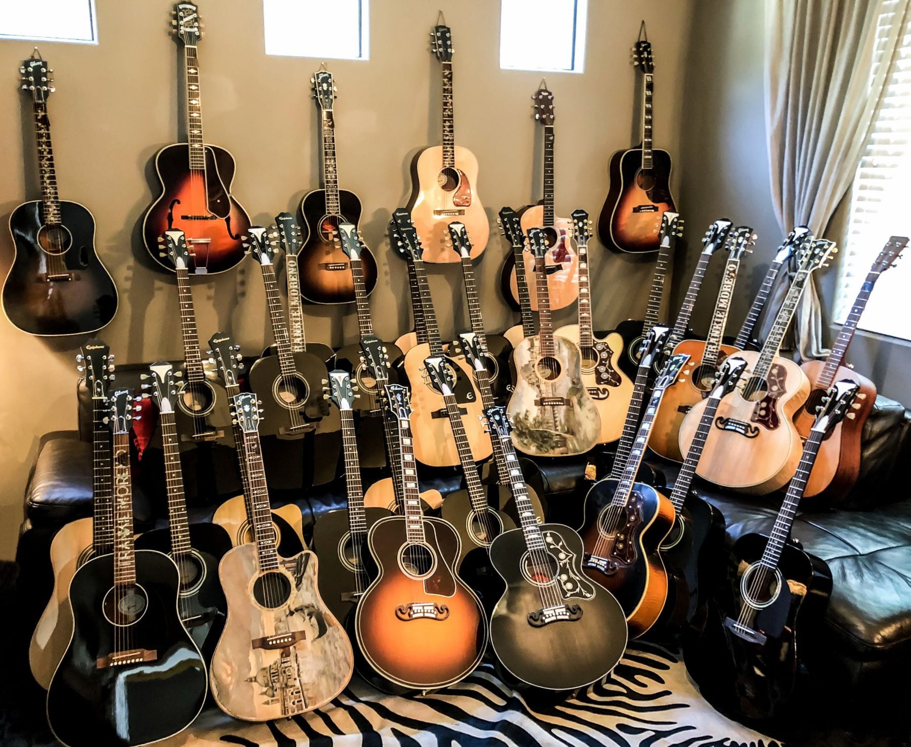 A collection of over 30 acoustic guitars, some hanging on a wall, others resting on a black leather couch.