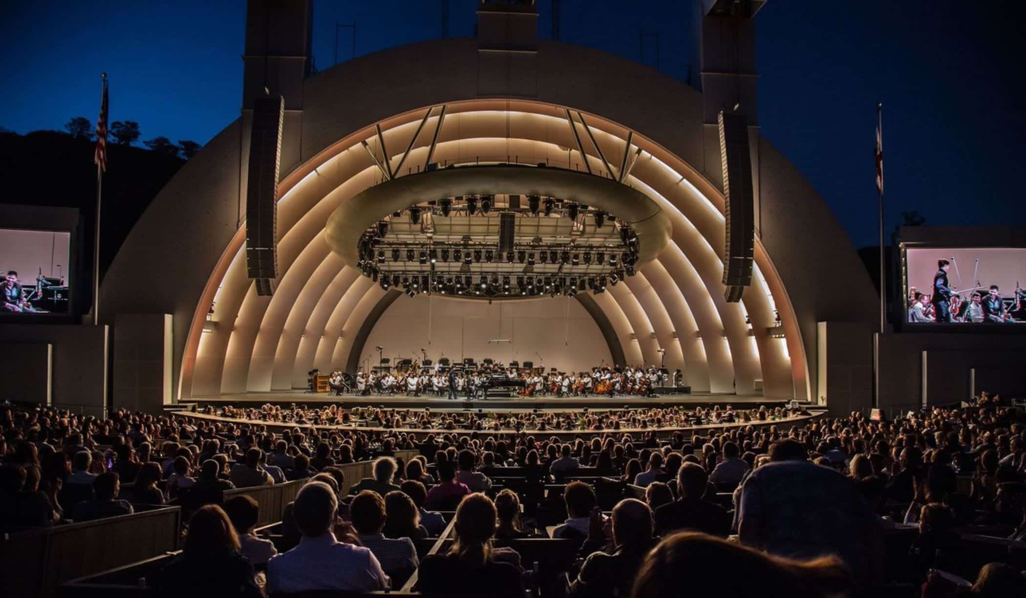 A large outdoor amphitheater at dusk, with an orchestra performing on a brightly lit stage to a full audience.