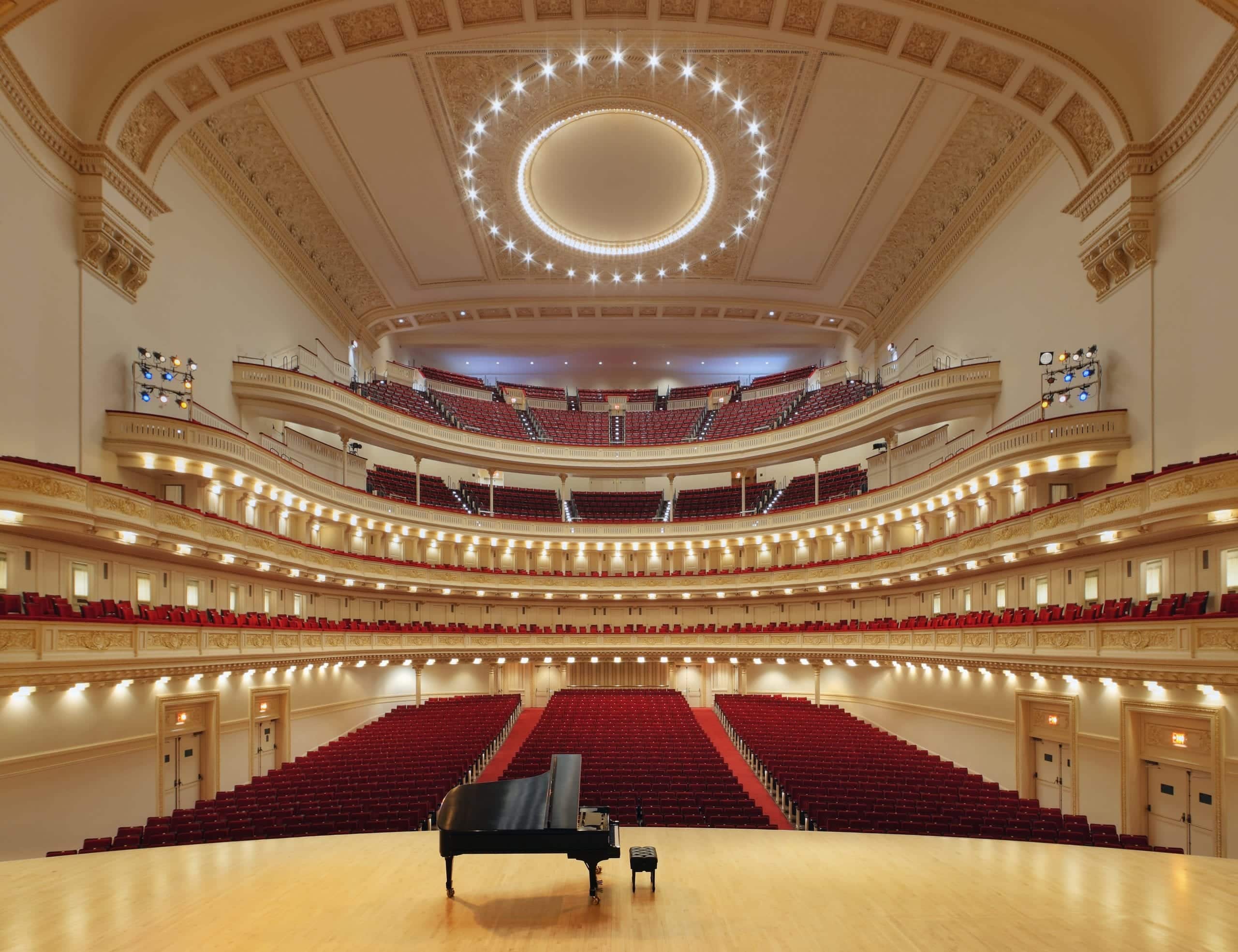 A grand concert hall with a black piano on a wooden stage, surrounded by rows of empty red seats.