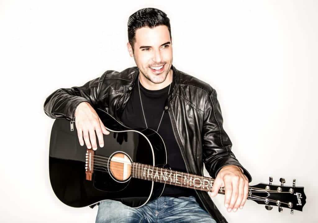 A smiling man in a black leather jacket and jeans holds a black acoustic guitar with "FRANKIE MORE" on the fretboard.