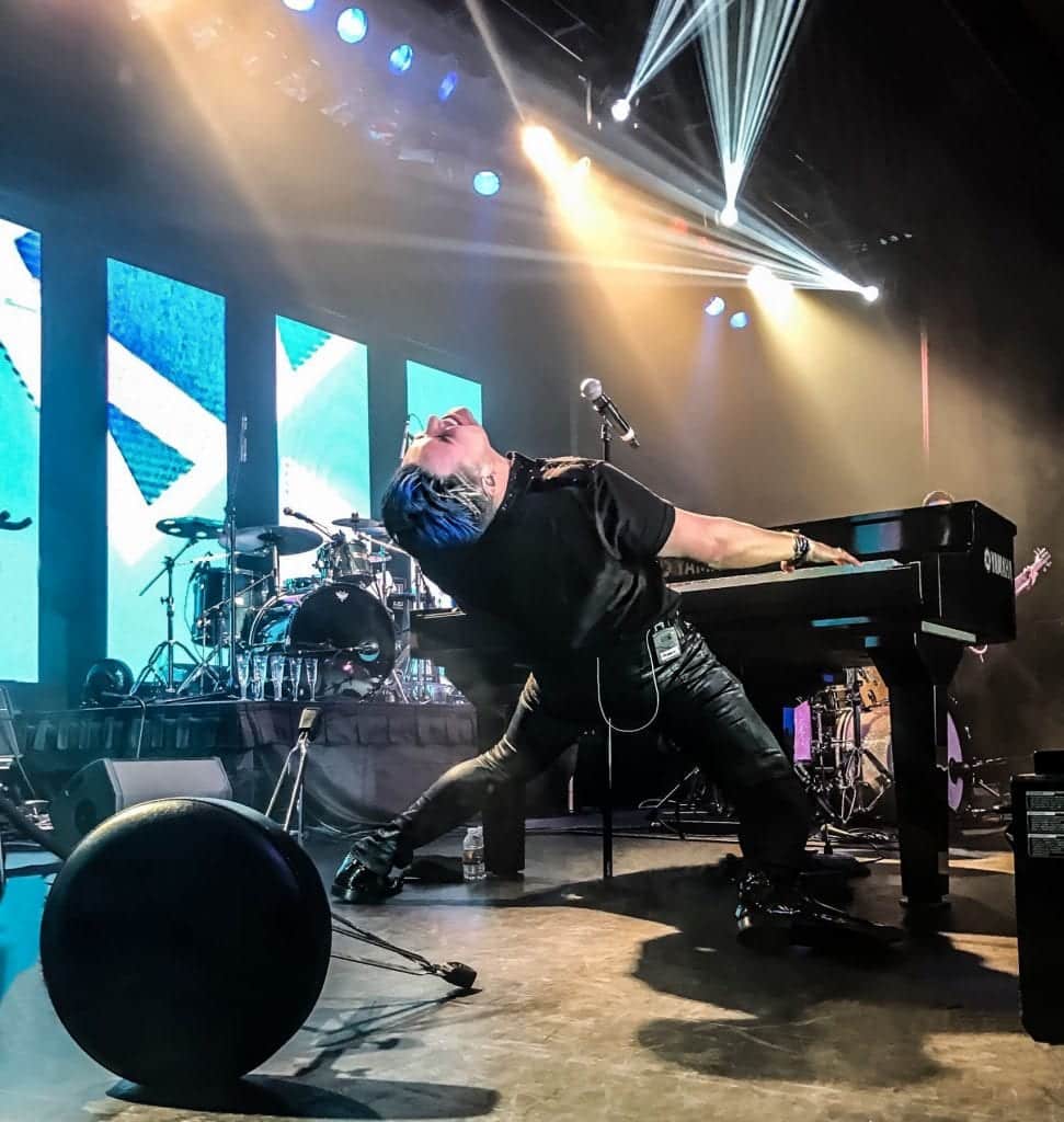 A musician with blue hair and a black shirt leans back dramatically over a "YAMAHA" keyboard on stage.