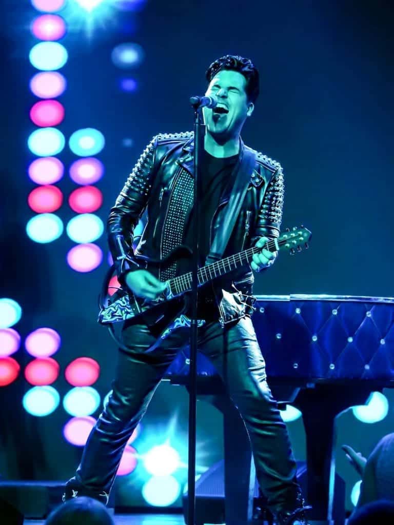 A male musician in a studded leather jacket and pants sings into a microphone while playing a guitar.