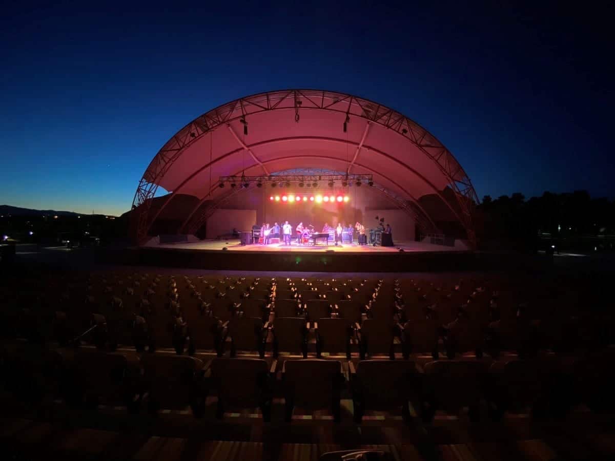 A band performs on a brightly lit stage under a large arched canopy at an outdoor amphitheater at dusk.