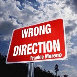 A red rectangular sign on a pole reads "WRONG DIRECTION Frankie Moreno" against a cloudy sky and dark trees.
